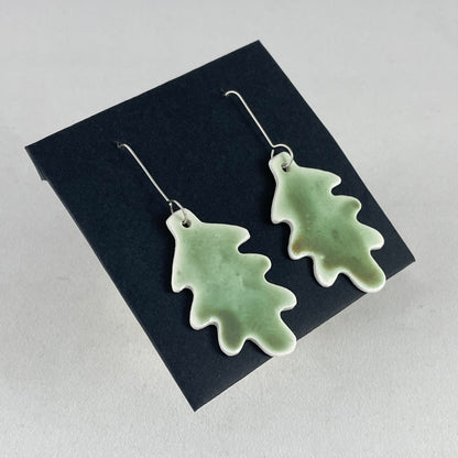 A pair of handmade porcelain earrings in oak leaf shape and warm green glaze. Photographed on a black background. On sterling silver earrings hooks. Side view