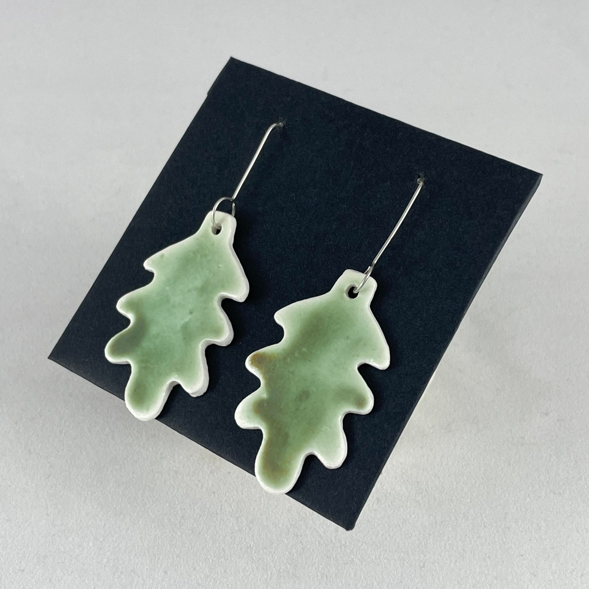 A pair of handmade porcelain earrings in oak leaf shape and warm green glaze. Photographed on a black background. On sterling silver earrings hooks. Side view