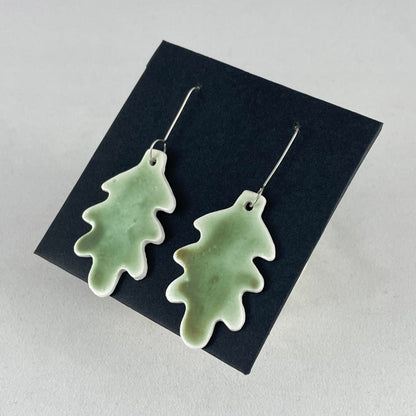 A pair of handmade porcelain earrings in oak leaf shape and warm green glaze. Photographed on a black background. On sterling silver earrings hooks. Side view