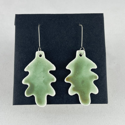 A pair of handmade porcelain earrings in oak leaf shape and warm green glaze. Photographed on a black background. On sterling silver earrings hooks.