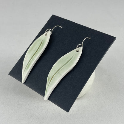 A pair of hand formed porcelain earrings in organic gum leaf shape. Photographed on a black background. On sterling silver earrings hooks. Side view