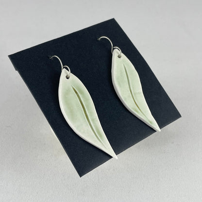 A pair of hand formed porcelain earrings in organic gum leaf shape. Photographed on a black background. On sterling silver earrings hooks. Side view